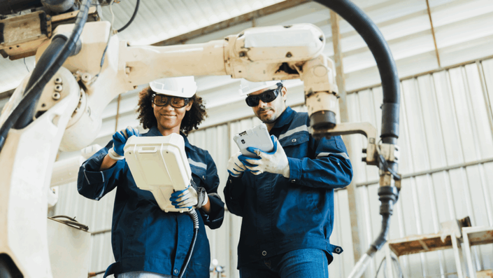 Two engineers in safety gear operate a large industrial robot arm inside a factory, illustrating collaboration between humans and machines in the workplace driven by AI and Automation.